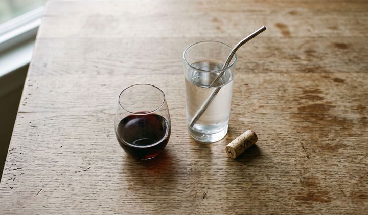 Halb gefülltes Rotweinglas neben einem Glas Wasser und einem Korken auf einem Holztisch im Abendlicht
