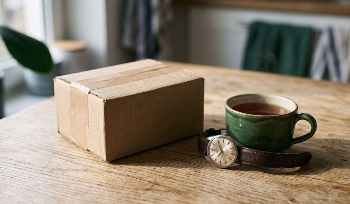 Ungeöffnetes Päckchen neben einer analogen Armbanduhr und einer Tasse Tee auf einem Holztisch im Morgenlicht