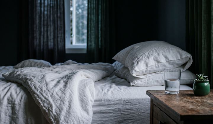 Ungemachtes Bett mit weißer Bettwäsche im dunklen Schlafzimmer, fahles Morgenlicht durchs Fenster, Wasserglas auf dem Nachttisch