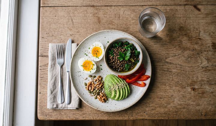 Teller mit Eiern, Linsen, Blattgrün, Avocado und Nüssen auf einem Holztisch im Morgenlicht am Fenster