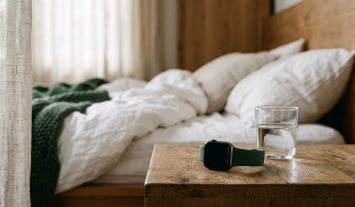 Smartwatch mit dunklem Display neben einem Wasserglas auf dem Nachttisch, weiches Morgenlicht durch einen Leinenvorhang