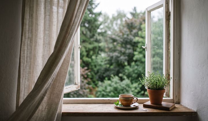 Offenes Fenster am Morgen, wehender Leinenvorhang, Teetasse und Kräutertopf auf der Fensterbank, draußen grüne Bäume im Licht