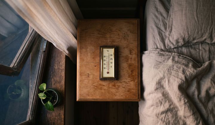 Thermometer auf einem Nachttisch neben zerwühltem Leinenbett, Fenster gekippt, warmes Abendlicht geht in blaue Nacht über