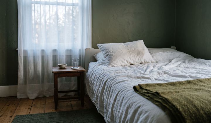 Ruhiges Bett mit Leinenbezug und Wolldecke im weichen, kühlen Morgenlicht, daneben ein Wasserglas auf dem Nachttisch.